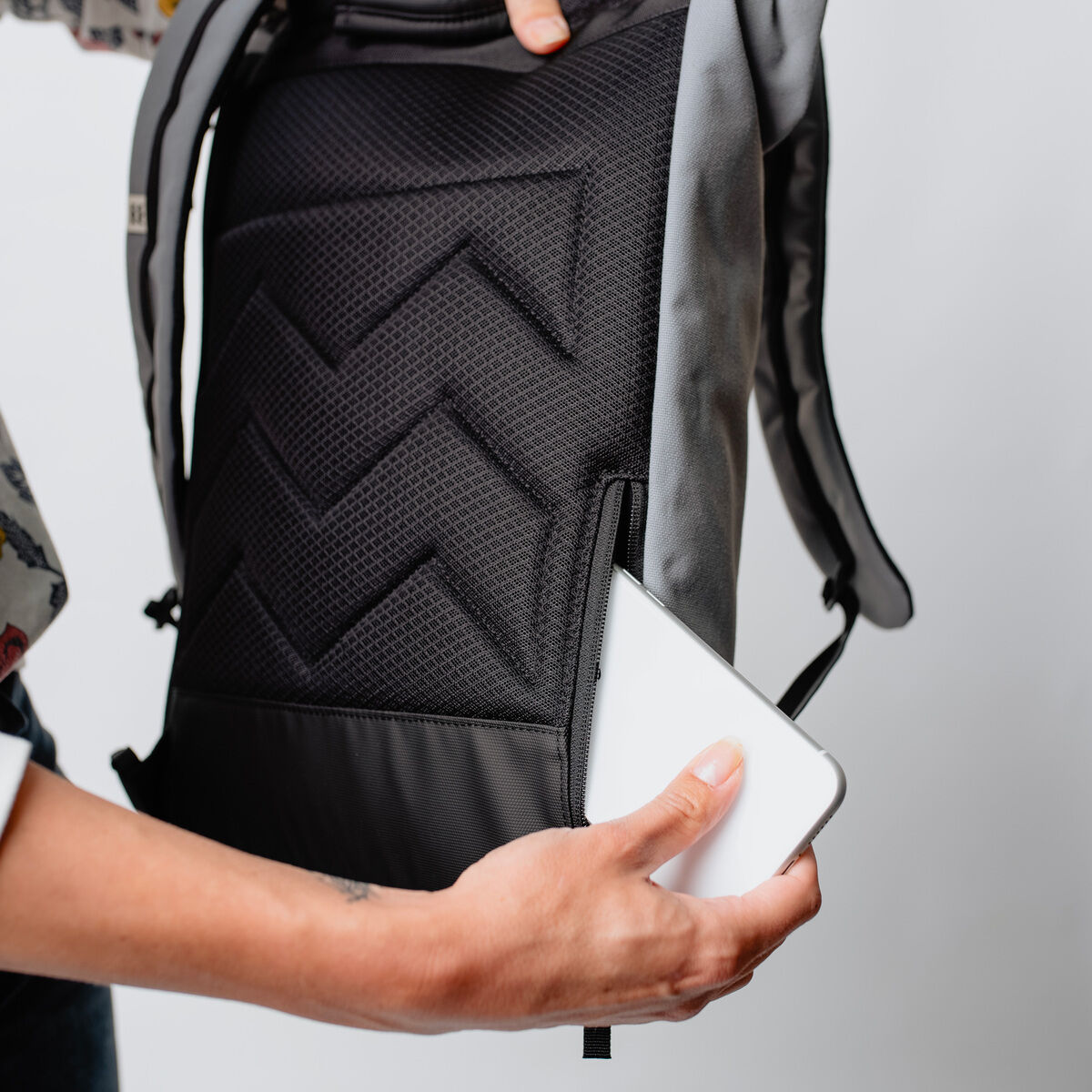 Nahaufnahme einer Person, die einen Rucksack »Wanaka« trägt. Die Person hält ein weißes Smartphone, das aus einer Reißverschlusstasche des Rucksacks herausragt. Der Rucksack ist hauptsächlich schwarz und grau mit einer strukturierten Oberfläche und Chevron-Muster.