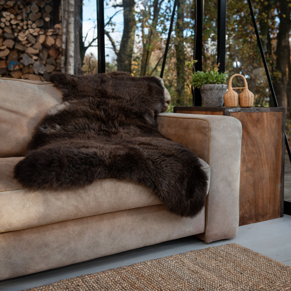 Ein dunkelbraunes Schafsfell mit Lederschlaufe liegt dekorativ auf einer beigen Couch in einem modernen Wohnzimmer mit großen Fenstern und Holzdeko. Ein Beistelltisch mit Pflanzen und gemütlicher Ausstrahlung steht daneben.