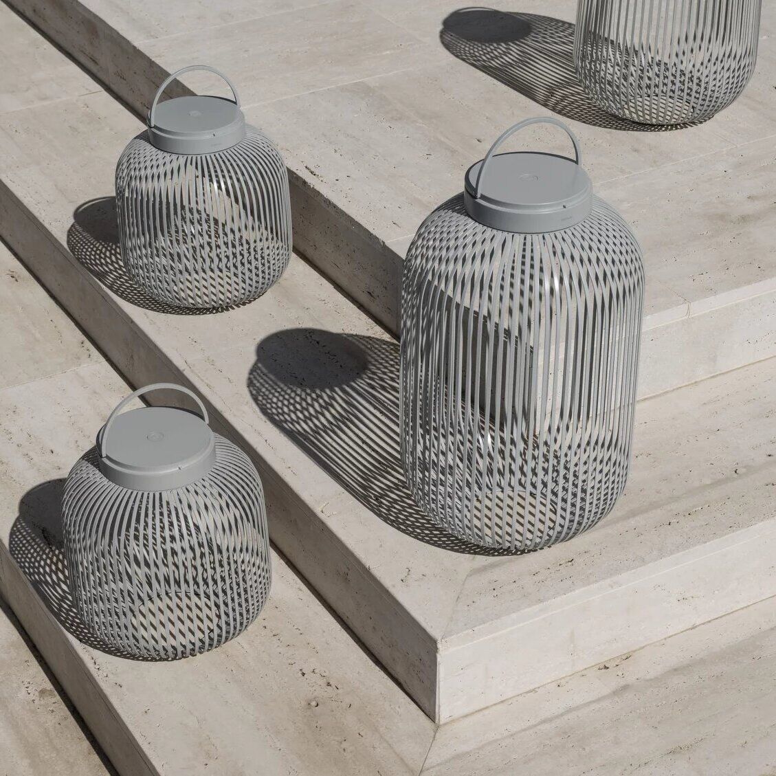 Eine Gruppe von modernen, tragbaren LED Laternen der Größe M in grau, die auf Natursteinstufen im Freien platziert sind. Die Laternen haben ein zylindrisches Design mit seitlichen Schlitzen und einem Henkel an der Oberseite.