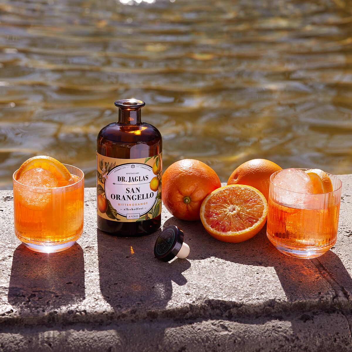Auf einem Stein am Ufer stehen eine Flasche San Orangello alkoholfreier Bitterorangen-Aperitif von Dr. Jaglas, zwei Tumblergläser mit orangenen Getränken und Orangenscheiben sowie frische, aufgeschnittene Orangen. Im Hintergrund ist leicht verschwommenes Wasser zu erkennen.