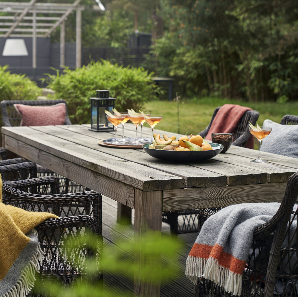 Eine gemütliche Gartenszene mit einem Holztisch, umgeben von Stühlen, die mit Decken überzogen sind. Auf dem Tisch stehen Gläser mit Getränken und eine Schale mit Obst. Im Hintergrund sind Grünpflanzen und ein Pool zu sehen.
