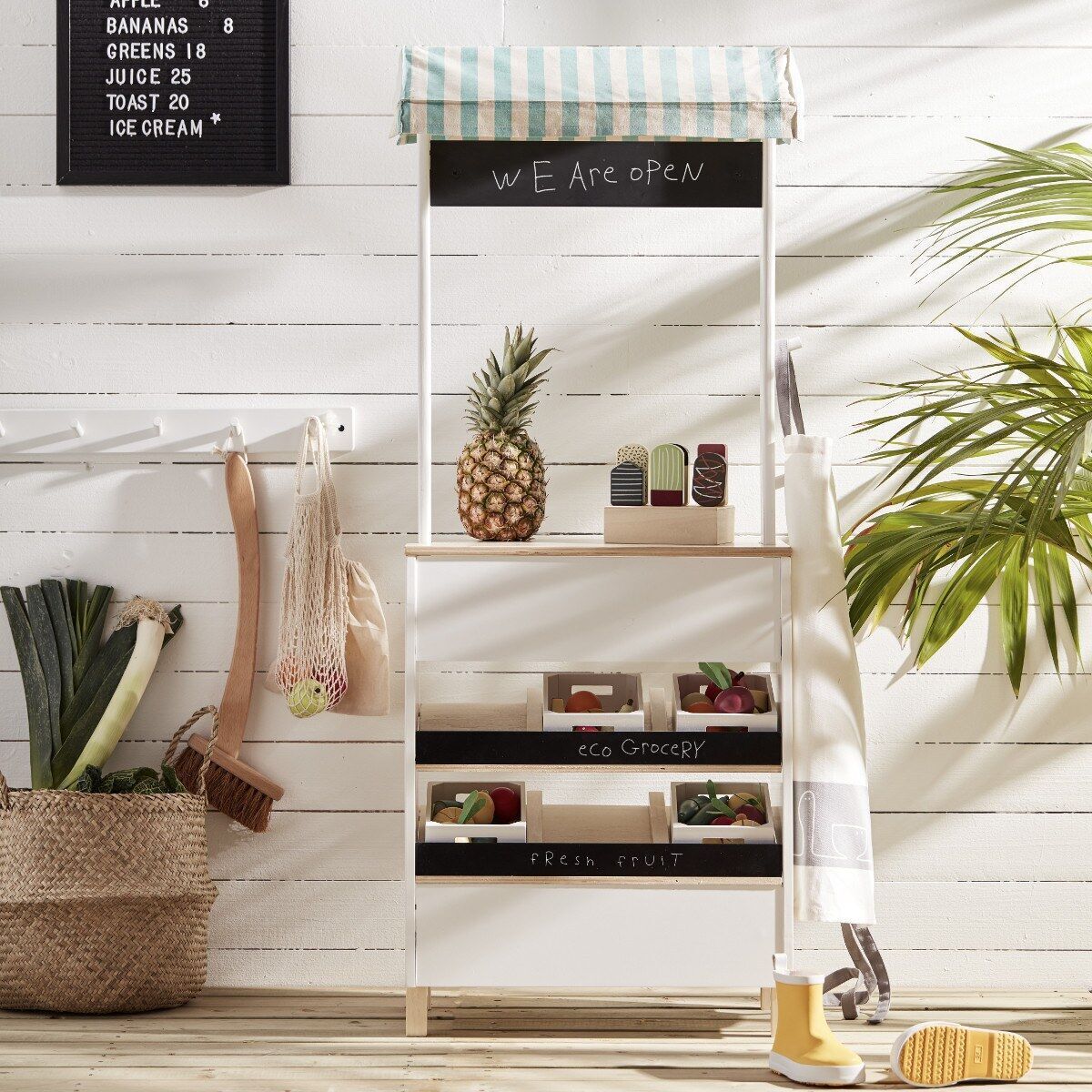 Ein Marktstand für Kinder aus Holz. Der Stand hat ein weiß-grün gestreiftes Dach und eine Tafel mit der Aufschrift 'We Are Open'. Darunter befinden sich Spielzeugfrüchte und -gemüse in kleinen Kisten und ein Einkaufskorb. Daneben stehen ein Besen und eine Gießkanne. Der Marktstand ist dekoriert mit Pflanzen.