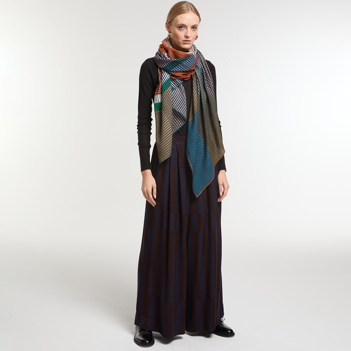 Frau trägt einen gemusterten Wollschal in den Farben Braun, Blau, Grün und Orange sowie ein langes, dunkles Kleid und einen schwarzen Pullover, aufgenommen vor neutralem Hintergrund. Das Outfit wirkt warm und modisch.