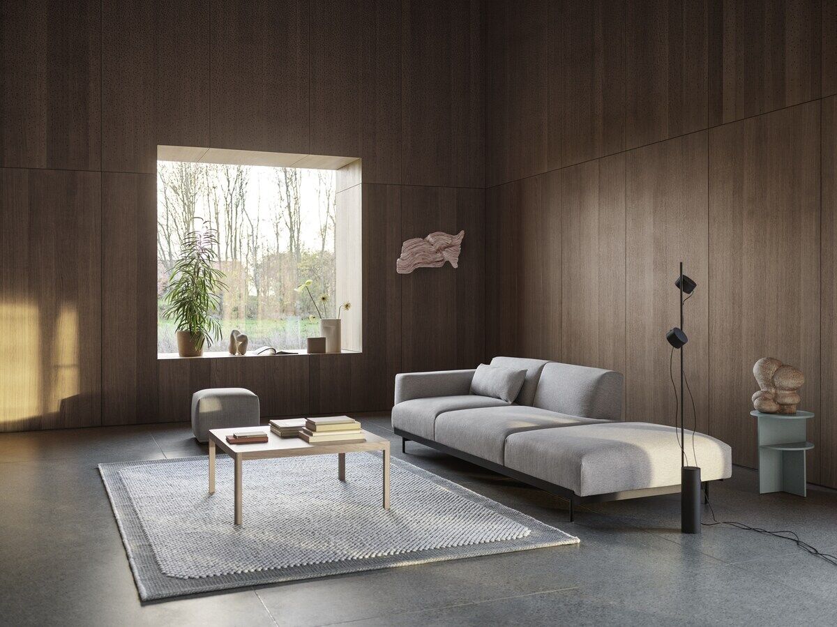 Modernes Wohnzimmer mit einer grauen Couch, einem Couchtisch aus Holz, einem grauen Teppich und einer schwarzen Bodenlampe »Post«. Im Hintergrund ein großes Fenster mit Pflanzen, das viel Tageslicht hereinlässt. Die Wände sind holzvertäfelt.