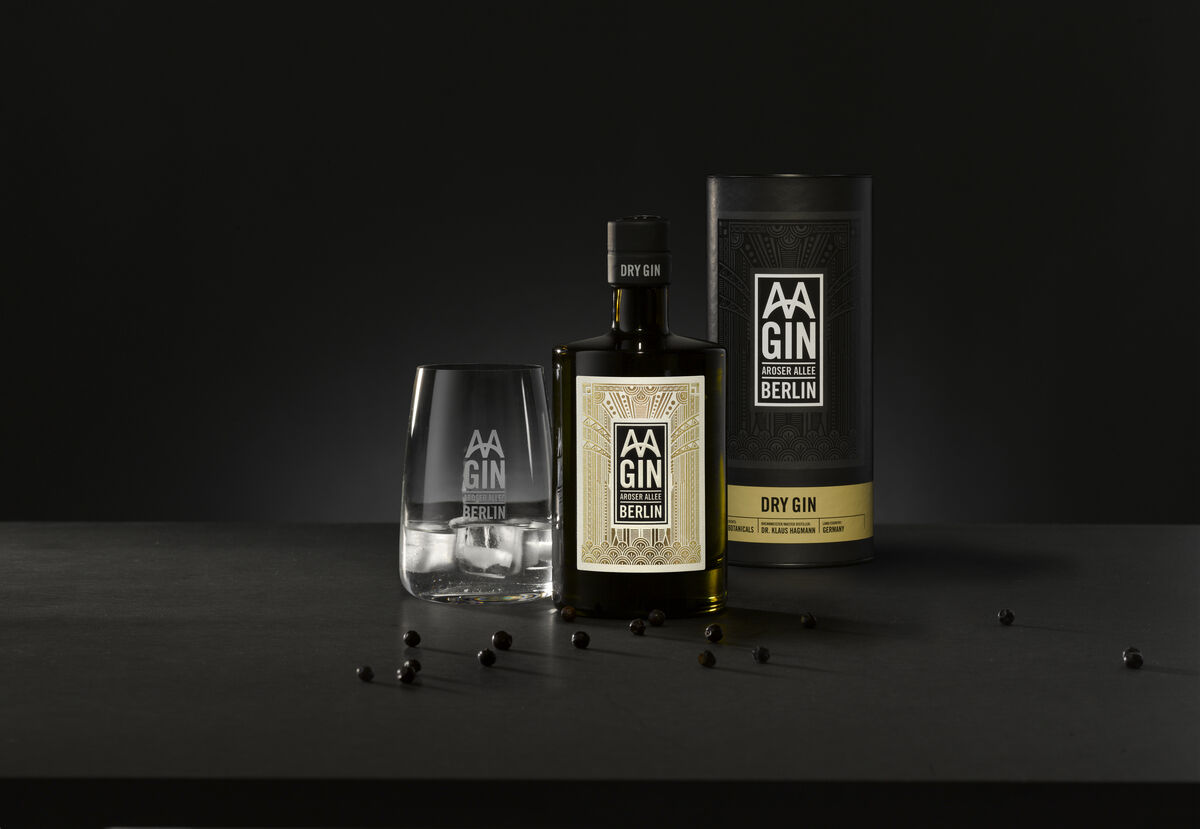 Ein Set aus »AAGIN« Dry Gin und einem dazugehörigen Glas. Der Gin wird in einer eleganten Flasche und einer Tube-Verpackung präsentiert. Einige Wacholderbeeren sind um das Set verstreut. Das Bild hat einen dunklen Hintergrund.