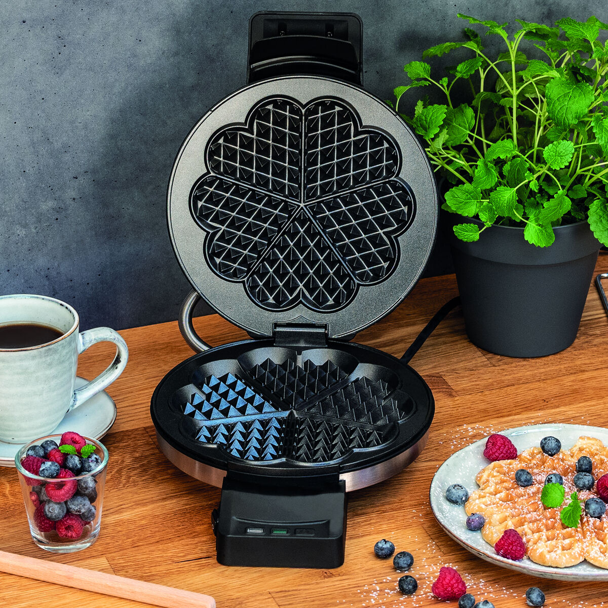 Das Bild zeigt ein Waffeleisen der Marke Cloer-1898 auf einem Holztisch. Neben dem Gerät befinden sich eine Tasse Kaffee, ein Glas mit Beeren und frische, gepflückte Waffeln mit Obst und Puderzucker garniert. Im Hintergrund steht eine grüne Pflanze.