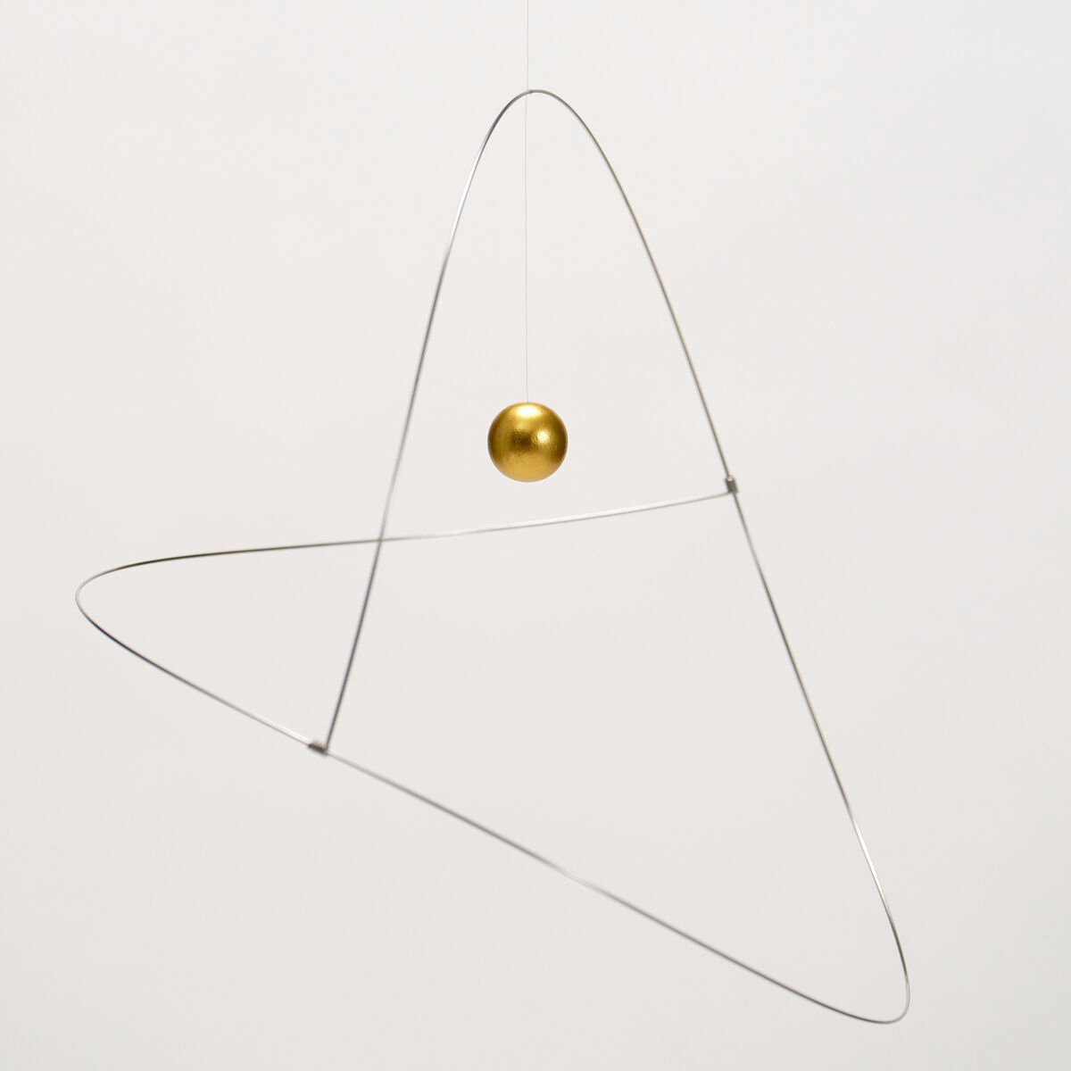 Das Bild zeigt das Mobile "Metamorphosis, Golden Planet" von Flensted Hanging Art: Eine schwebende goldene Kugel ist von zwei filigranen silbernen Drahtschlaufen umrahmt, die geometrisch und minimalistisch an dünnem Faden vor weißem Hintergrund hängen.