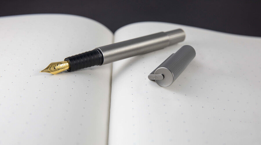 Füllfederhalter mit goldener Feder und grauem Metallkörper liegt auf einem aufgeschlagenen Notizbuch mit Punktraster, daneben die Kappe