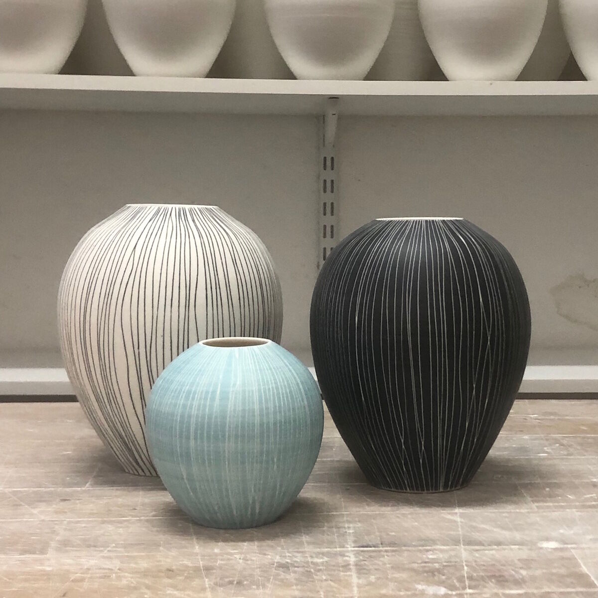 Drei Vasen in unterschiedlichen Größen und Farben auf einem Tisch. Die Vasen sind in den Farben Weiß, Schwarz und Hellblau und haben feine, vertikale Linien in einer Sgraffito-Technik.