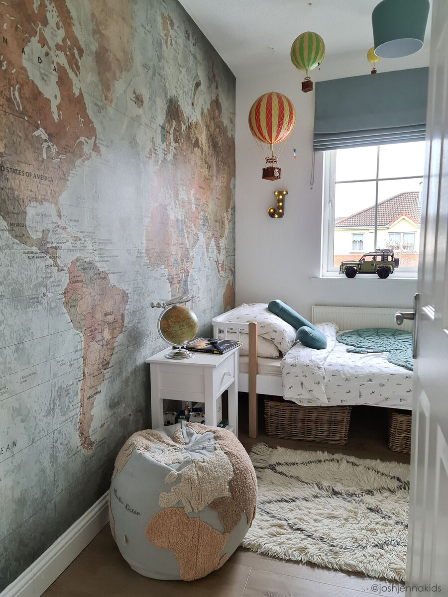 Gemütliches Kinderzimmer mit Weltkarten-Wanddekoration. Ein Sitzkissen in Form eines Weltkarten-Poufs steht neben einem kleinen weißen Tisch. Farbenfrohe Heißluftballons hängen an der Decke.