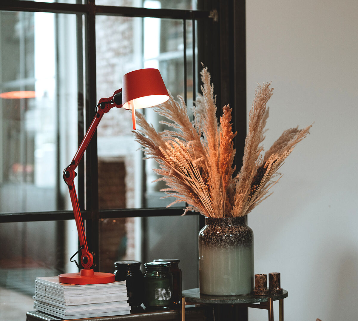Eine rote Schreibtischlampe »Bolt« steht auf einem Tisch vor einem Fenster. Daneben eine Vase mit getrockneten Gräsern und einige Bücher.