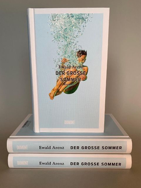 Das Bild zeigt drei Exemplare des Buches 'Ewald Arenz: Der große Sommer'. Ein Buch steht aufrecht, während die anderen beiden flach übereinander gestapelt sind. Auf dem Buchcover ist ein Junge im Wasser zu sehen.