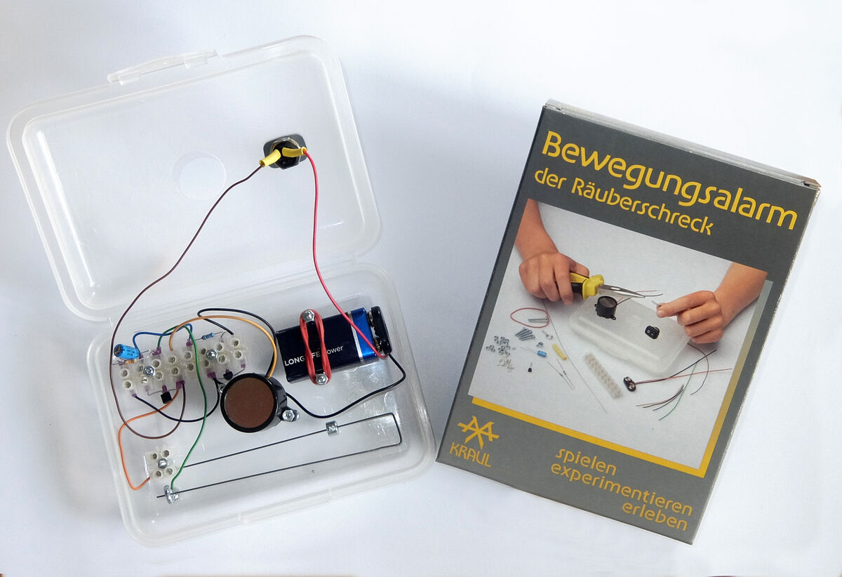 Der »Bewegungsalarm« Experimentierkasten von Kraul zeigt eine geöffnete Box mit Elektroteilen und Kabeln. Daneben steht eine Verpackung mit dem Bild des montierten Alarms und dem Text »Bewegungsalarm der Räuberschreck«. Der Kasten enthält diverse Bauteile, die für den Aufbau eines Bewegungsalarms notwendig sind.