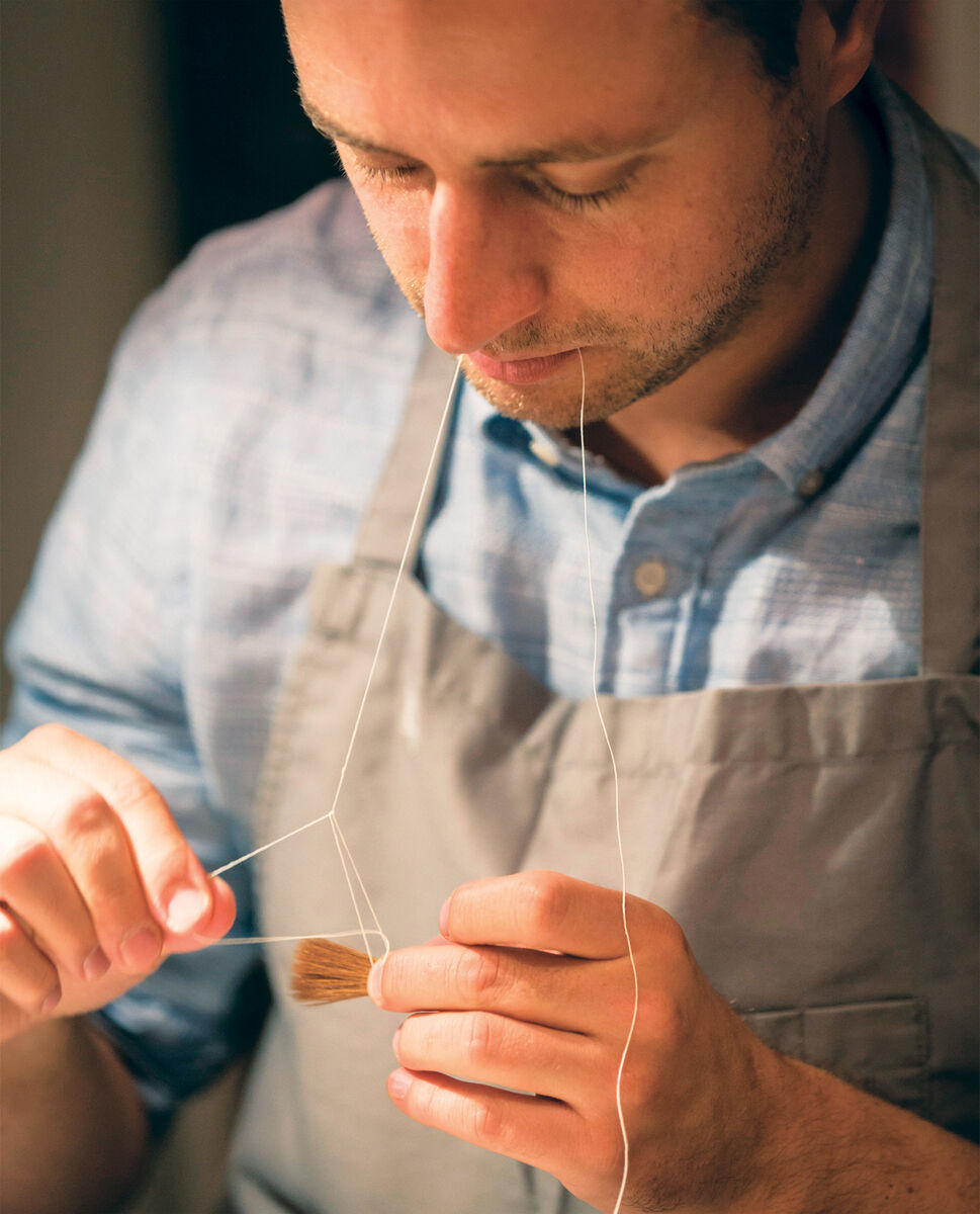 Ein Mann in einem grauen Kittel hält einen traditionellen Aquarellpinsel mit Ethergraf®-Metallspitze und einem Fadenwerkzeug in der Hand. Er konzentriert sich auf seine handwerkliche Arbeit. Der Hintergrund ist unscharf.
