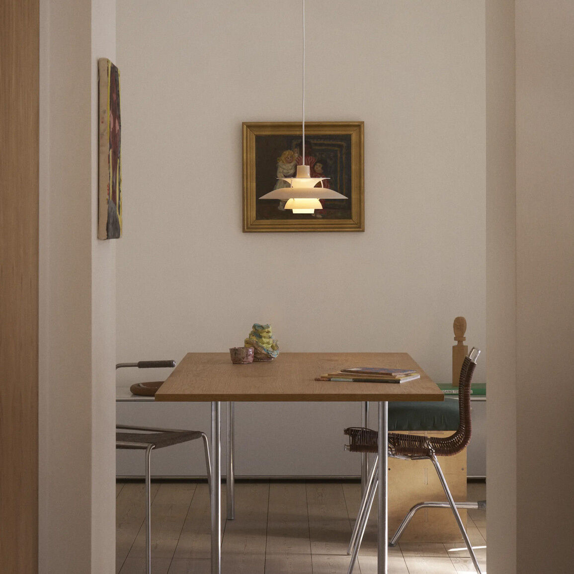 Im Zentrum hängt die beige-opale Hängeleuchte »PH 5 320« von Louis Poulsen über einem modernen, hellen Holztisch. Zwei Stühle, dezente Wandfarben, ein Gemälde und harmonische Lichtwirkung schaffen eine ruhige, elegante Wohnatmosphäre.