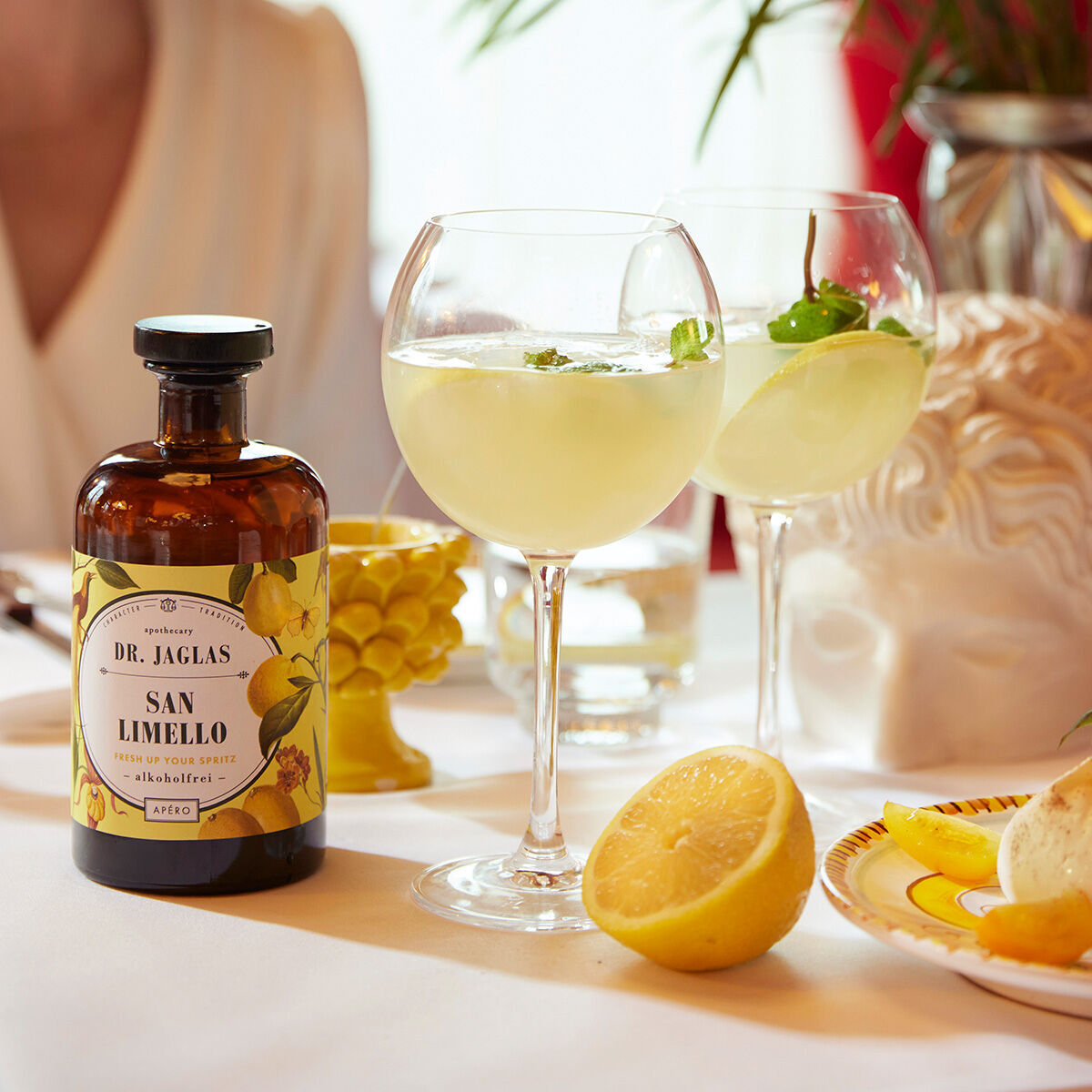 Eine Flasche alkoholfreier Aperitif »San Limello« von Dr. Jaglas steht auf einem Tisch neben zwei gefüllten Gläsern, einer halben Zitrone und dekorativen Elementen.