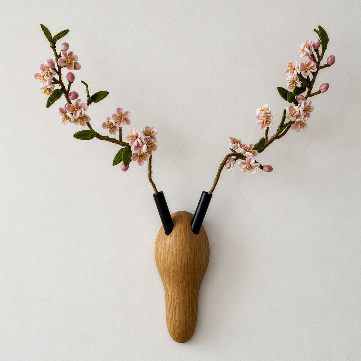 An einer mattweißen Wand hängt die Wanddeko »RESPECT«: ein schlanker, tropfenförmiger Sockel aus warmem braunem Eschenholz mit zwei schwarzen Röhrchen. Darin stecken verzweigte Zweige mit rosa Blüten, Knospen und dunkelgrünen Blättern, die wie ein stilisiertes Geweih nach oben wachsen.
