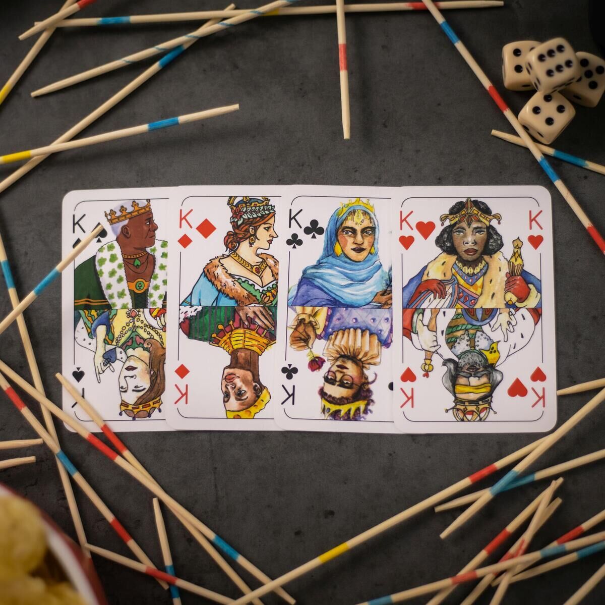 Spielkarten im Skat-Stil liegen auf einem Tisch, umgeben von Mikado-Stäben und Würfeln.