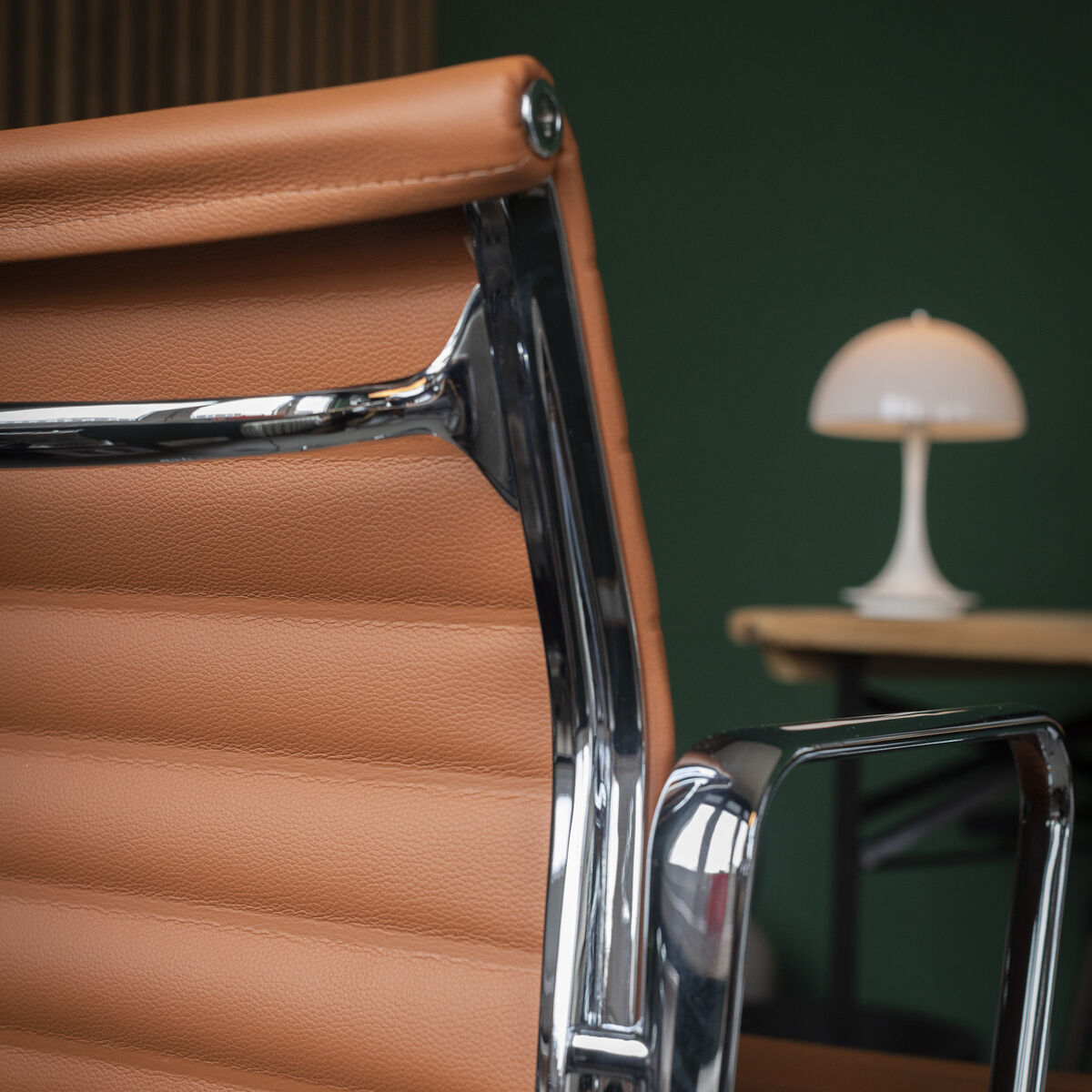 Detailansicht des VITRA Alu Chair EA 118 in cognacfarbenem Leder mit verchromtem Aluminiumgestell, im Hintergrund ein grüner Raum, ein heller Schreibtisch und eine kleine weiße Tischlampe auf heller Holzplatte. Ambiente wirkt modern und hochwertig.