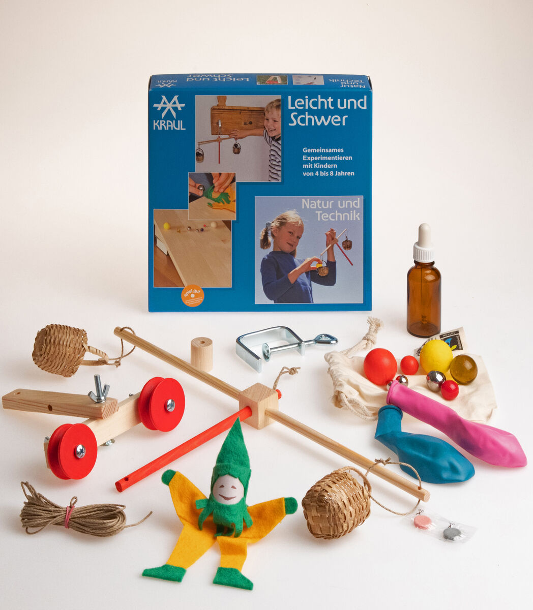 Ein Experimentierkasten namens »leicht und schwer« von Kraul mit verschiedenen Teilen wie Holzbauteilen, einem Seil, Rädern, einer Pipette, verschiedenen Bällen und einem grünen Puppenclown im Vordergrund. Auf der Verpackung sind Bilder von Kindern zu sehen, die mit dem Set experimentieren. Die Farben des Sets sind vielseitig, einschließlich Blau, Rot, Grün, Gelb, Braun und Weiß.