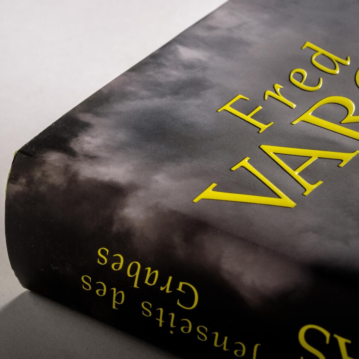 Nahaufnahme des Buchcovers von Fred Vargas' 'Jenseits des Grabes'. Das Cover zeigt den Titel in gelber Schrift und eine düstere, wolkenverhangene Szenerie in Grautönen. Auf dem Cover befindet sich ein runder Aufkleber mit Text.