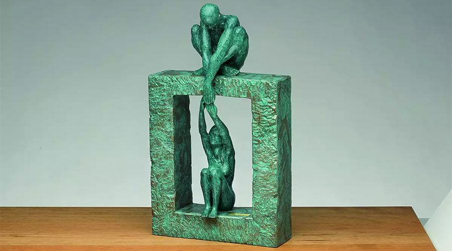 Bronzeskulptur zeigt zwei menschliche Figuren, eine sitzt auf einem rechteckigen Rahmen und hält die Hand einer anderen Figur, die im Rahmen hängt