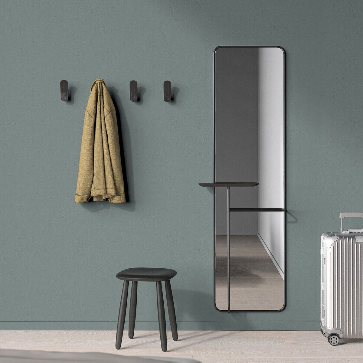 Spiegel »Concierge« an einer Wand mit grüner Tapete, darunter ein schwarzer Hocker und rechteckige, schwarze Wandhaken neben einem beigefarbenen Mantel, sowie ein silberner Koffer links von einem Bett mit grauer Bettwäsche.