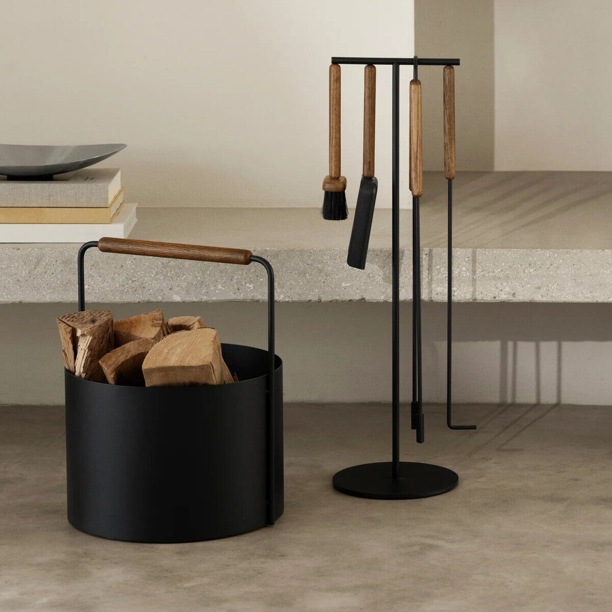 Abgebildet ist ein moderner Holzkorb aus schwarzem Metall mit Holzgriff, gefüllt mit Holzscheiten. Daneben steht ein elegantes Kaminbesteck mit Griffen aus hellem Holz. Die Szene spielt sich auf einer hellen, minimalistischen Steinfläche ab und vermittelt einen wohnlichen, skandinavischen Einrichtungsstil.