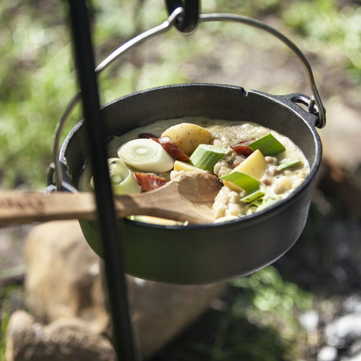 Ein Dutch Oven aus Gusseisen hängt über einem Lagerfeuer im Freien. Der Topf ist mit Gemüse und Fleisch gefüllt. Ein Holzkochlöffel ragt aus dem Topf heraus. Im Hintergrund sind Gras und Steine zu sehen.