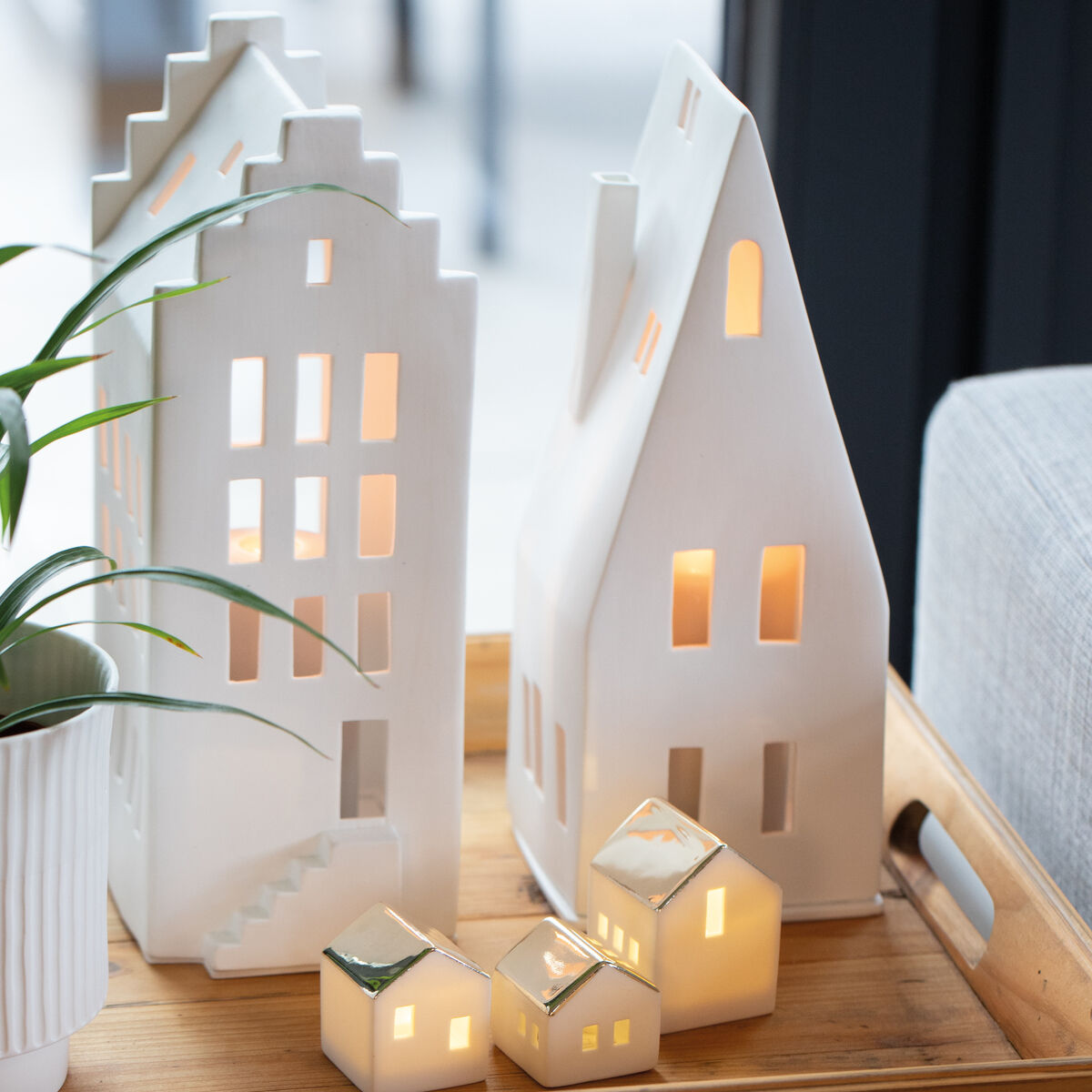 Zwei weiße keramische Lichthäuser auf einem Holztablett, mit warmem Licht beleuchtet, neben einer Pflanze und auf einem Holztisch platziert.
