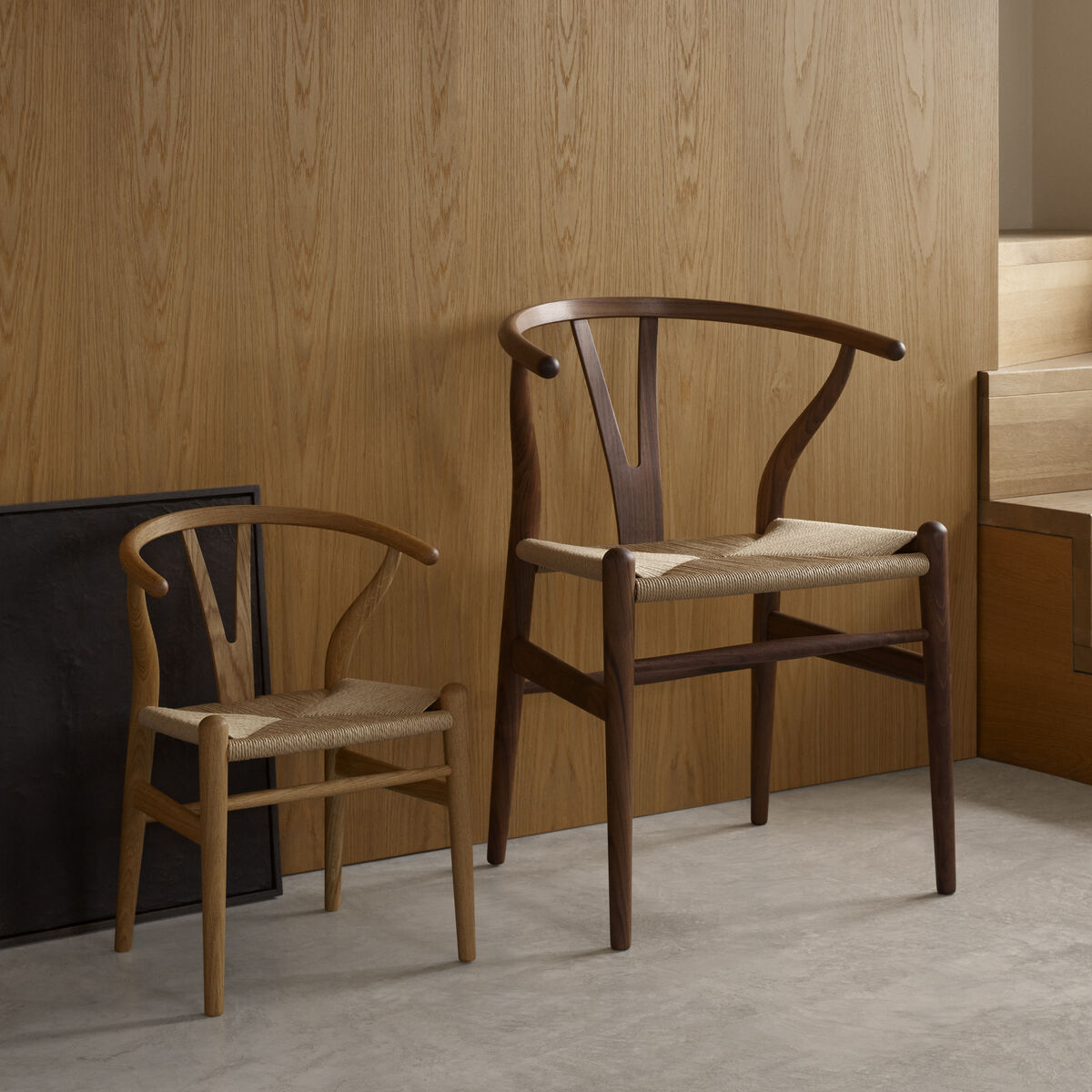 Ein Bild mit zwei Wishbone Chairs »CH24 Kinder«, einem kleinen und einem großen, aus Holz in einem minimalistischen Raum mit Holzwand und -treppe. Die Stühle haben eine natürliche Holzfarbe und Sitzflächen aus Flechtmaterial.