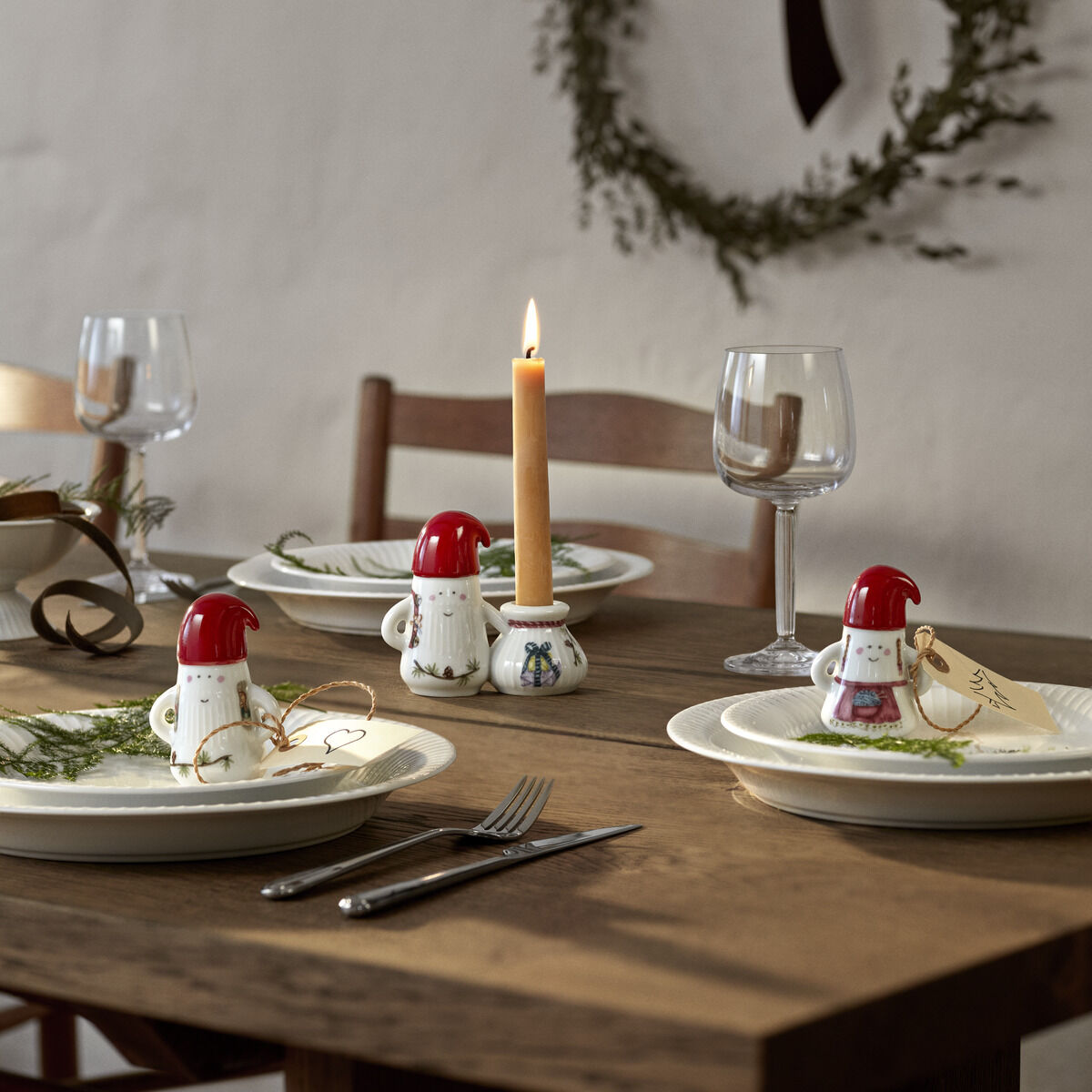 Keramik-Weihnachtsfrauen der Serie »Hammershøi« stehen als festliche Tischdekoration auf einem Holztisch, der für ein weihnachtliches Essen gedeckt ist. Im Hintergrund ist eine brennende Kerze und eine dekorative grüne Kranzgirlande an der Wand zu sehen.