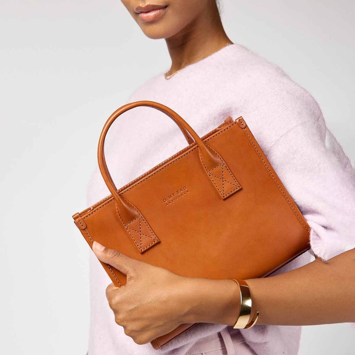 Eine modebewusste Frau trägt eine mittelgroße, braune Lederhandtasche mit zwei Henkeln. Die Tasche ist schlicht, elegant und hochwertig verarbeitet. Sie eignet sich für den Alltag, fürs Büro oder besondere Anlässe. Im Hintergrund trägt die Frau einen weichen, pastellfarbenen Pullover.