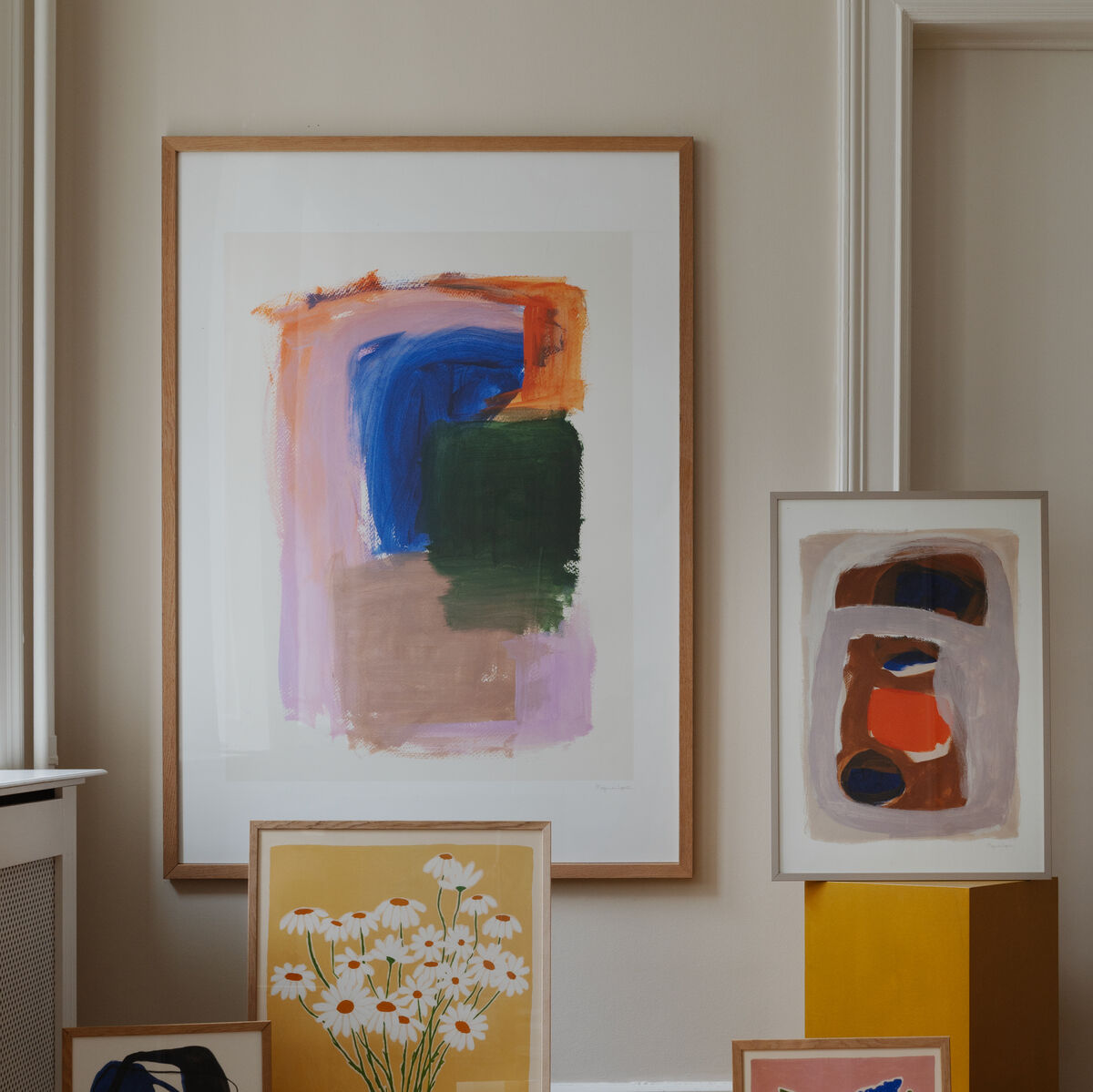 Eine Sammlung von gerahmten abstrakten Kunstwerken von Berit Mogensen Lopez, präsentiert auf einem Holzboden vor einer beigen Wand. Die Bilder zeigen kräftige Farben in Blau, Orange, Grün und enthalten floral inspirierte Motive. Perfekte dekorative Ergänzung für moderne Innenräume.