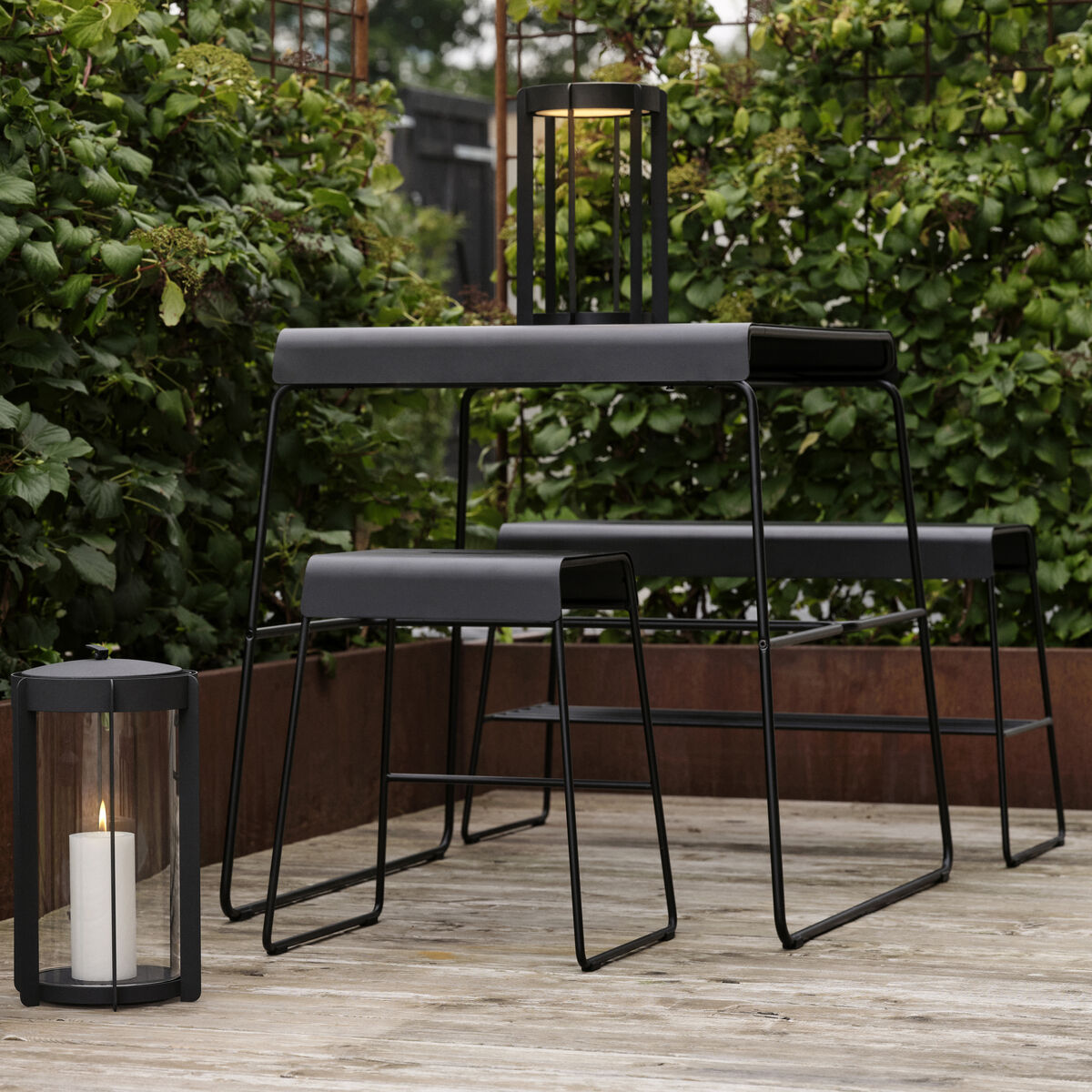 Ein moderner Outdoor-Schemel steht auf einer Holzterrasse, umgeben von grünen Sträuchern. Neben dem Schemel befinden sich zwei Laternen, in denen weiße Kerzen brennen. Ideal für Garten- oder Terrasseneinrichtungen.