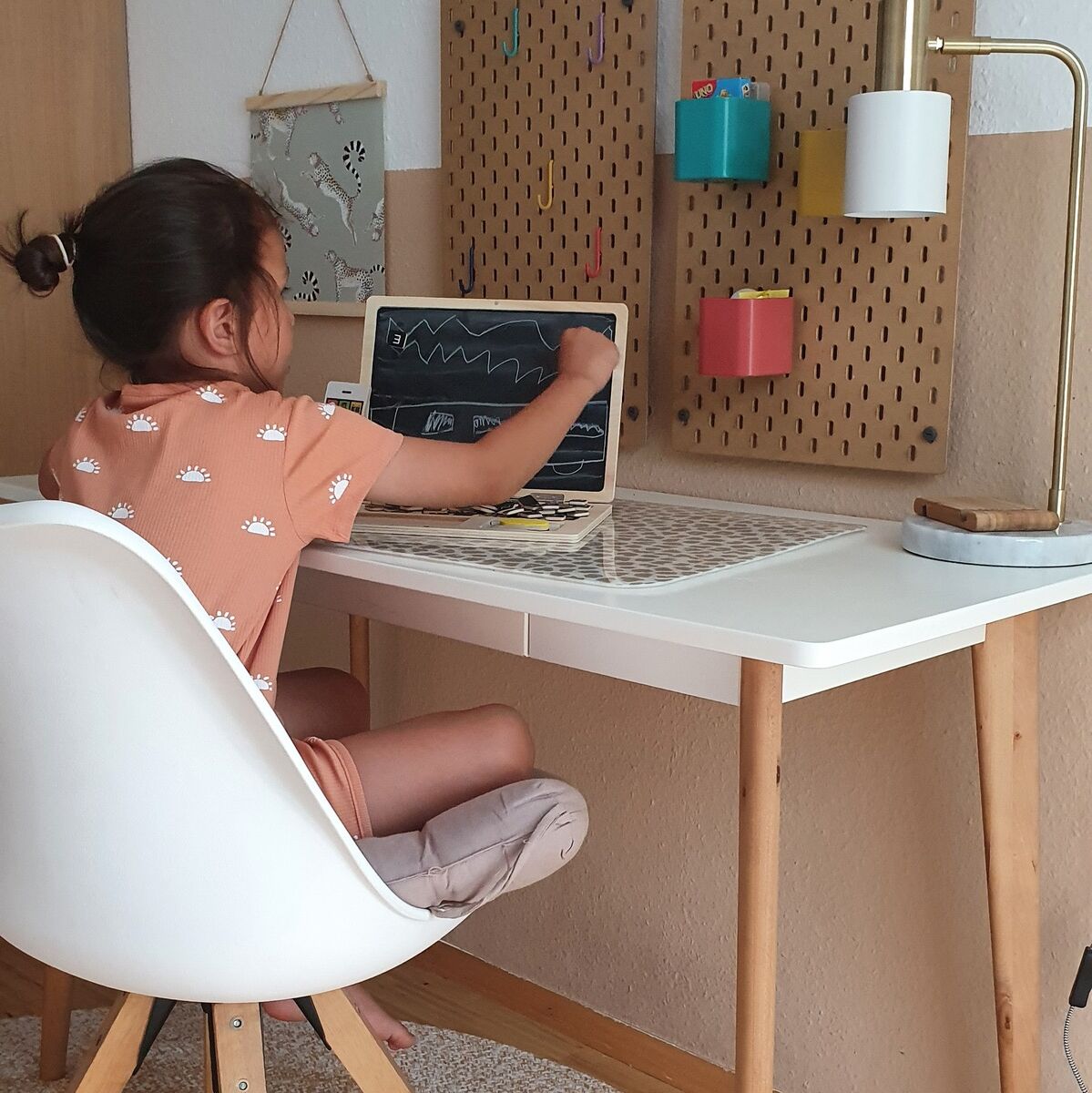 Ein Kind sitzt an einem Schreibtisch und spielt mit einem Holz Laptop mit Magnettafel. Im Hintergrund hängen an der Wand zwei Tafeln und eine Lampe steht auf dem Schreibtisch. Der Raum ist hell und gemütlich eingerichtet.