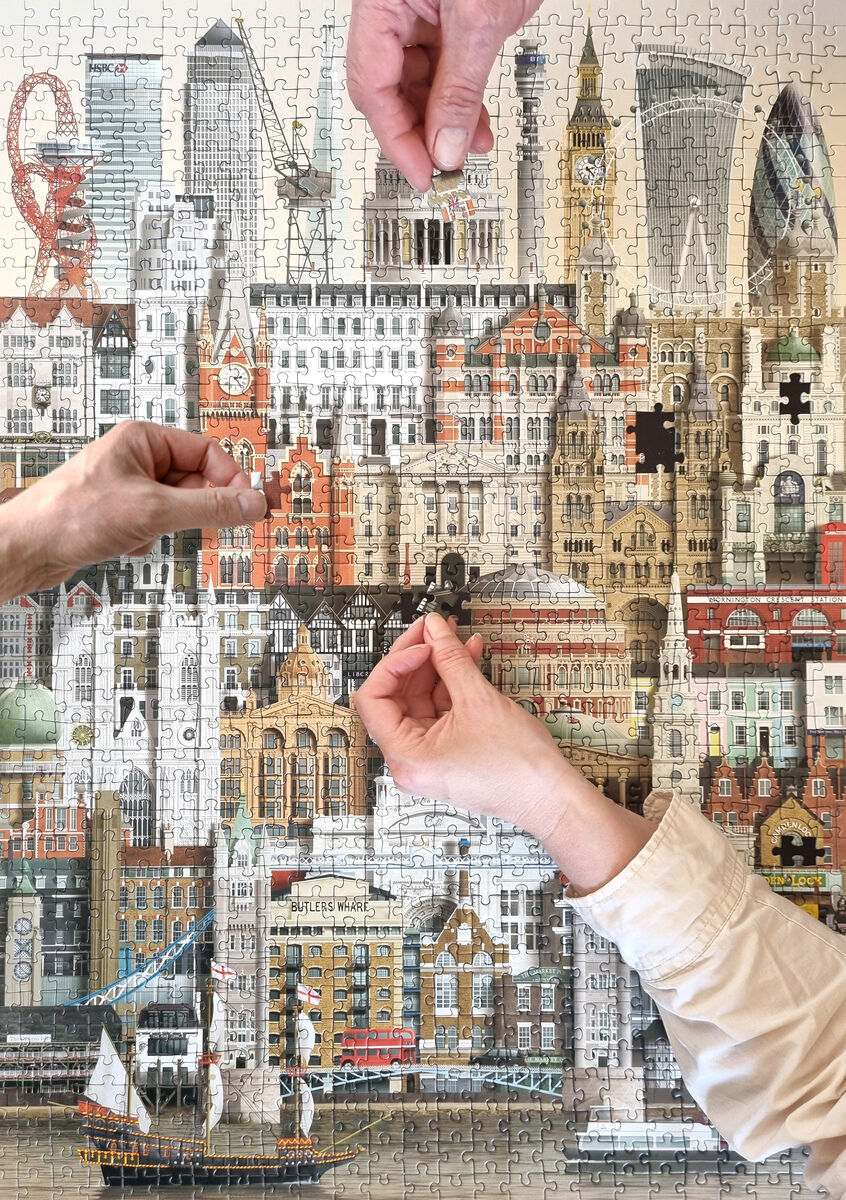 Ein fast fertiggestelltes Puzzle von London mit 1000 Teilen. Mehrere Hände setzen die letzten Puzzleteile ein. Sichtbar sind berühmte Sehenswürdigkeiten wie Big Ben und die Tower Bridge. Die Farben sind überwiegend Beige, Braun, Grau, Weiß, Schwarz und Rot.