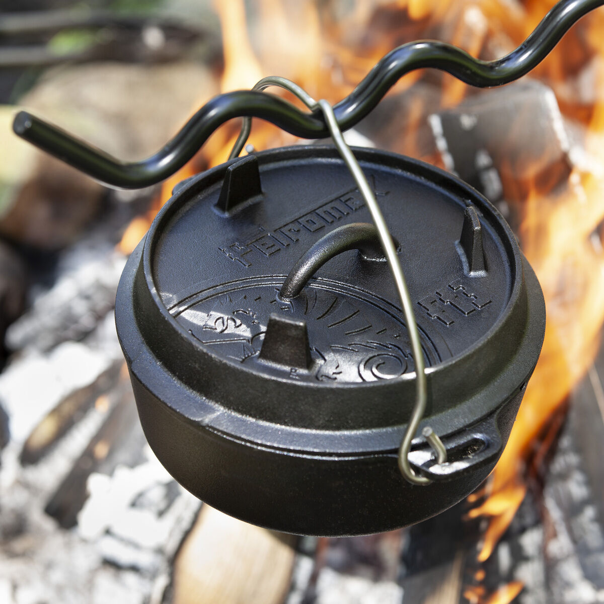Ein schwarzer Dutch Oven aus Gusseisen hängt über einer offenen Feuerstelle. Im Hintergrund sind Flammen und brennendes Holz zu sehen.