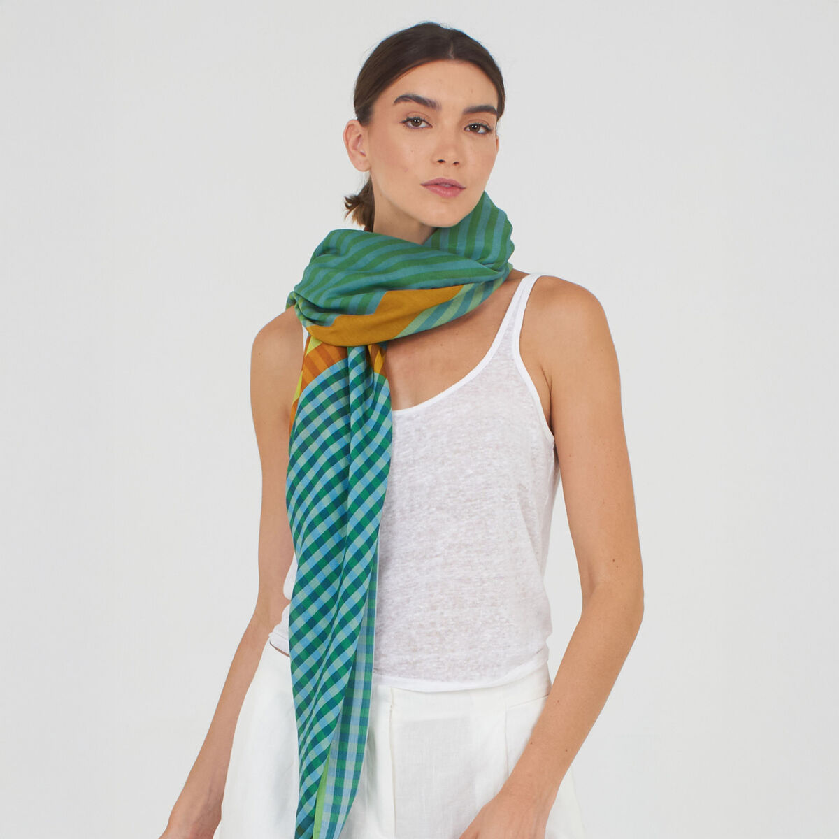 Frau mit locker um den Hals gewickeltem grünen Baumwoll-Seidenschal »Willa« von Shalls Barcelona; der Schal zeigt ein Streifen- und Karomuster in verschiedenen Grüntönen mit gelben und blauen Akzenten, kombiniert zu weißem Top und weißer Hose, vor hellem Hintergrund.