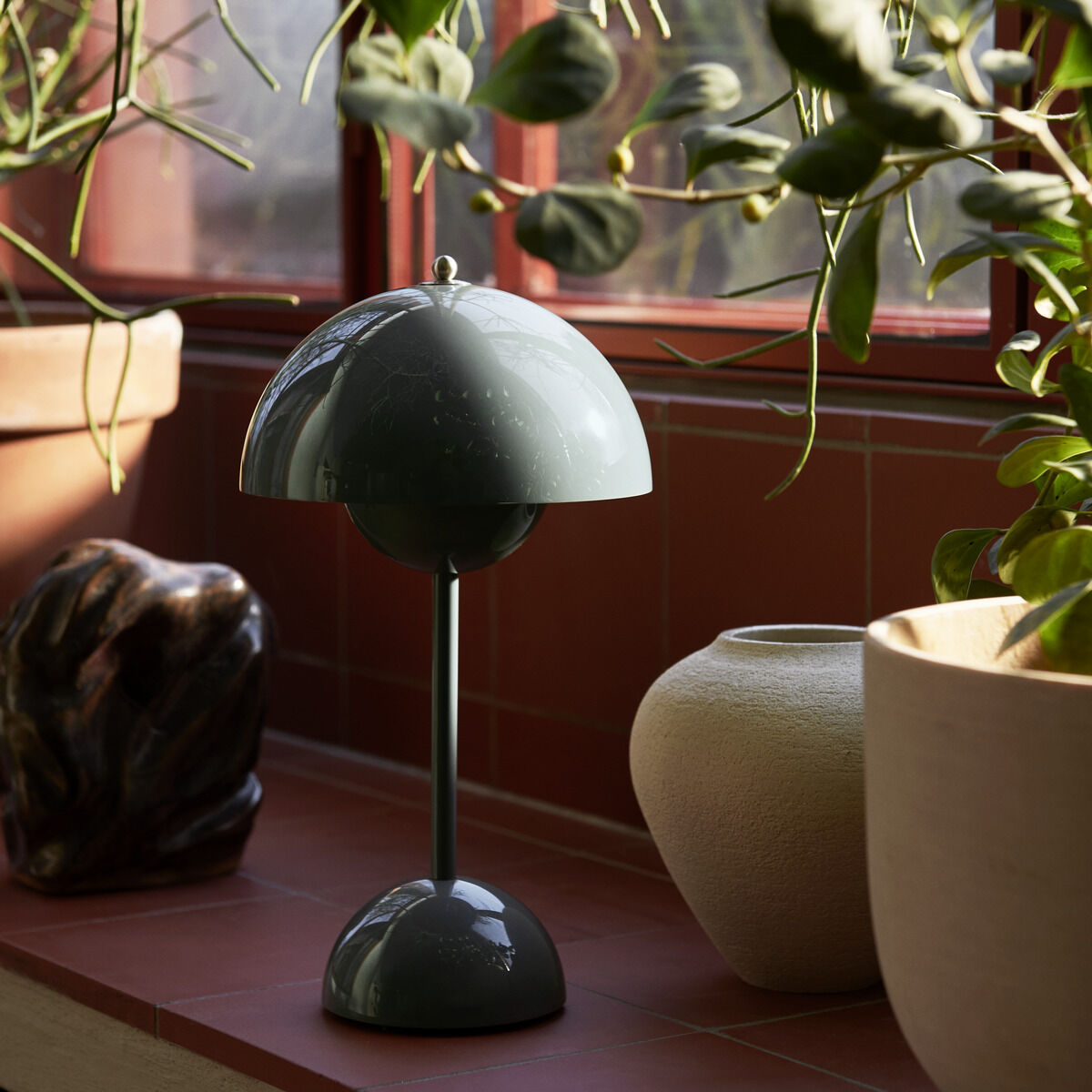 Die Flowerpot VP9 Tischleuchte Akku steht auf einer Fensterbank, umgeben von grünen Pflanzen. Die Lampe hat ein elegantes, skandinavisches Design in Grün und fügt sich harmonisch in die dezente rote und beige Umgebung ein.