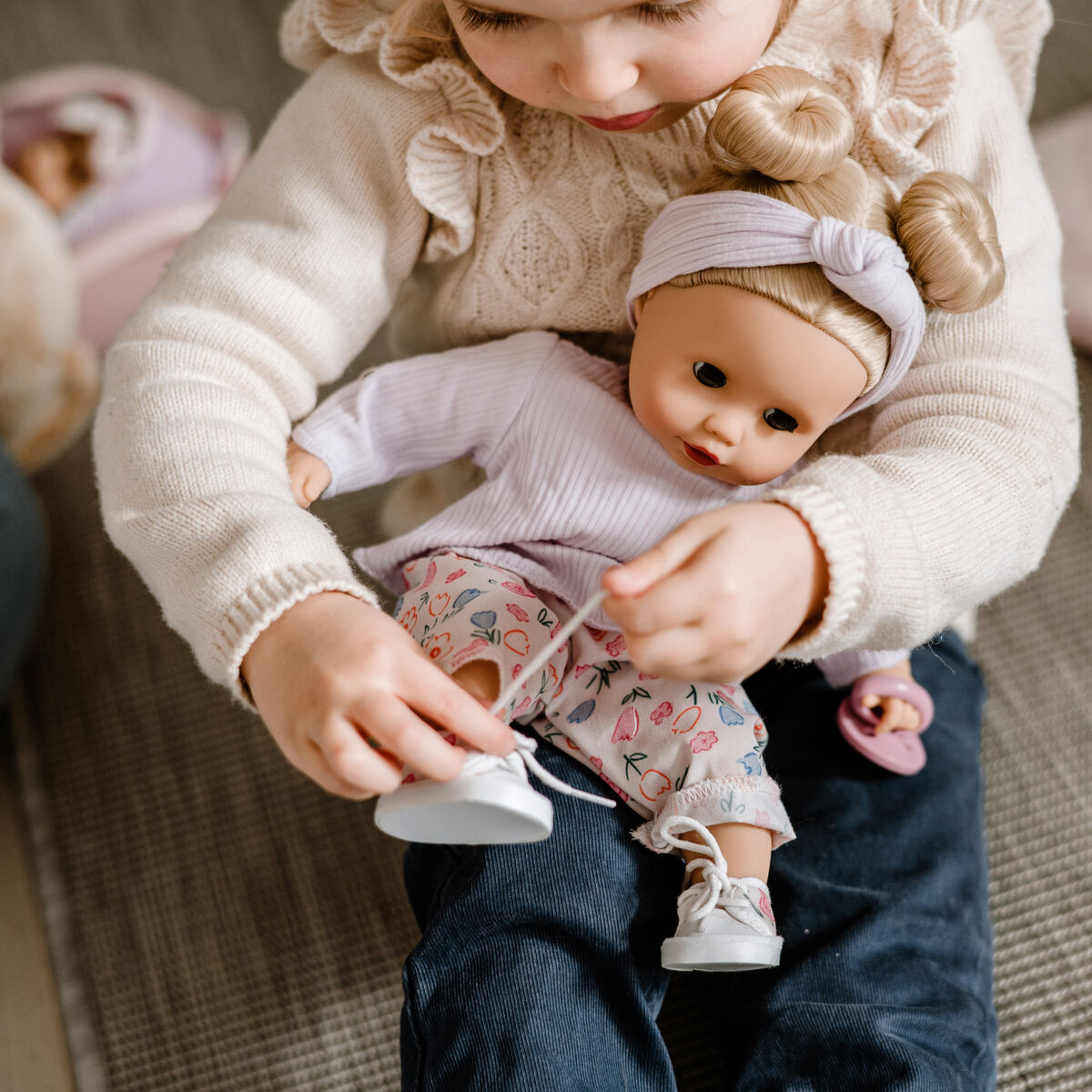 Ein kleines Kind spielt auf dem Boden mit einer Puppe namens Muffin BliBlaBlume, zieht ihr Schuhe an und hält sie liebevoll auf dem Schoß. Die Puppe trägt ein pastellfarbenes Outfit mit gemusterten Hosen und einem Stirnband. Die Szene zeigt ein harmonisches, kindgerechtes Spiel im Wohnraum.