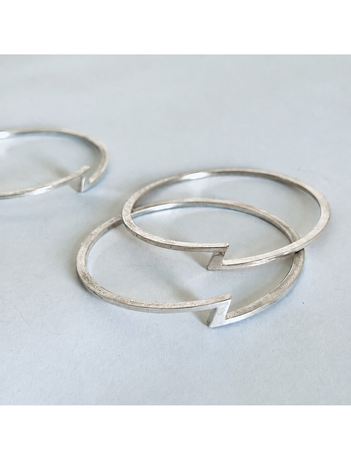 Zwei minimalistische Armreifen aus 925er Silber mit charakteristischem Zickzack-Detail liegen auf einer hellgrauen Oberfläche. Die Armreifen wirken modern und puristisch, das markante Z greift das Jubiläum der ZEIT auf. Die Silberoberfläche ist mattiert.