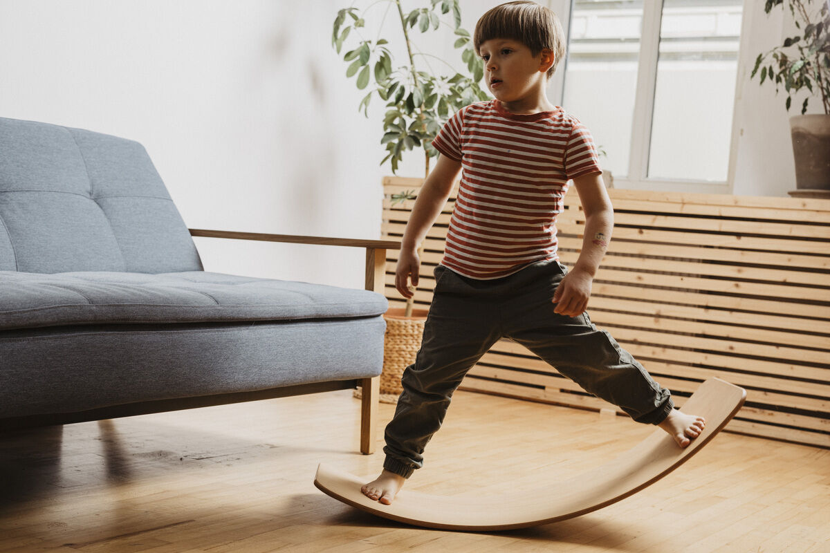 Ein Kind balanciert auf einem Balance Board aus Holz in einem Wohnzimmer mit blauer Couch, Holzboden und grünen Pflanzen.