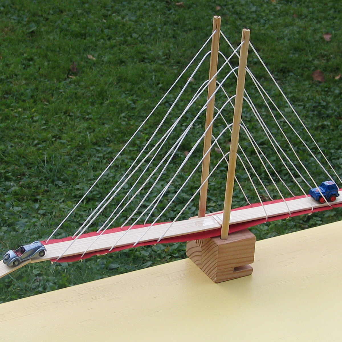 Auf dem Foto ist ein aufgebauter Bausatz einer Hängebrücke aus Holz zu sehen, der auf einer gelben Oberfläche steht. Über die Brücke fahren zwei kleine Modellautos. Im Hintergrund ist eine grüne Grasfläche erkennbar. Die Brücke enthält zahlreiche gespannte weiße Seile zur Stabilisierung und ein zentrales Brückenfundament.