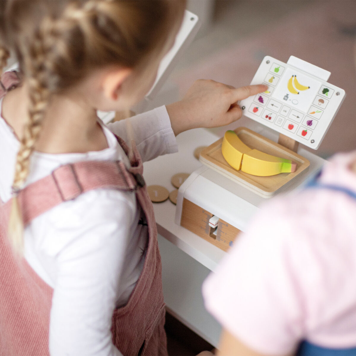 Zwei Kinder spielen mit einer hölzernen Spielzeugwaage von Waage »Fresh«. Das Bild zeigt eine detaillierte Nachbildung einer Waage sowie Spielzeugfrüchte und ein Display mit bunten Tasten und einer Banane.