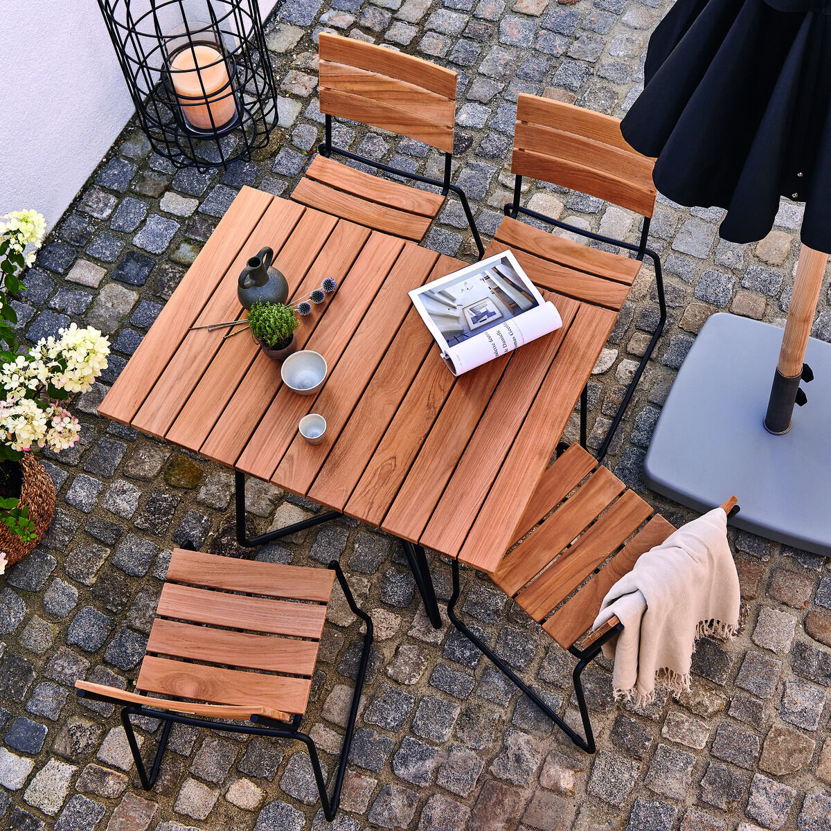 Ein klappbarer Holztisch aus Teakholz, umgeben von passenden Klappstühlen und dekoriert mit einer Laterne, Pflanzen und einem aufgeschlagenen Buch. Der Tisch steht auf einem steinernen Boden im Außenbereich.