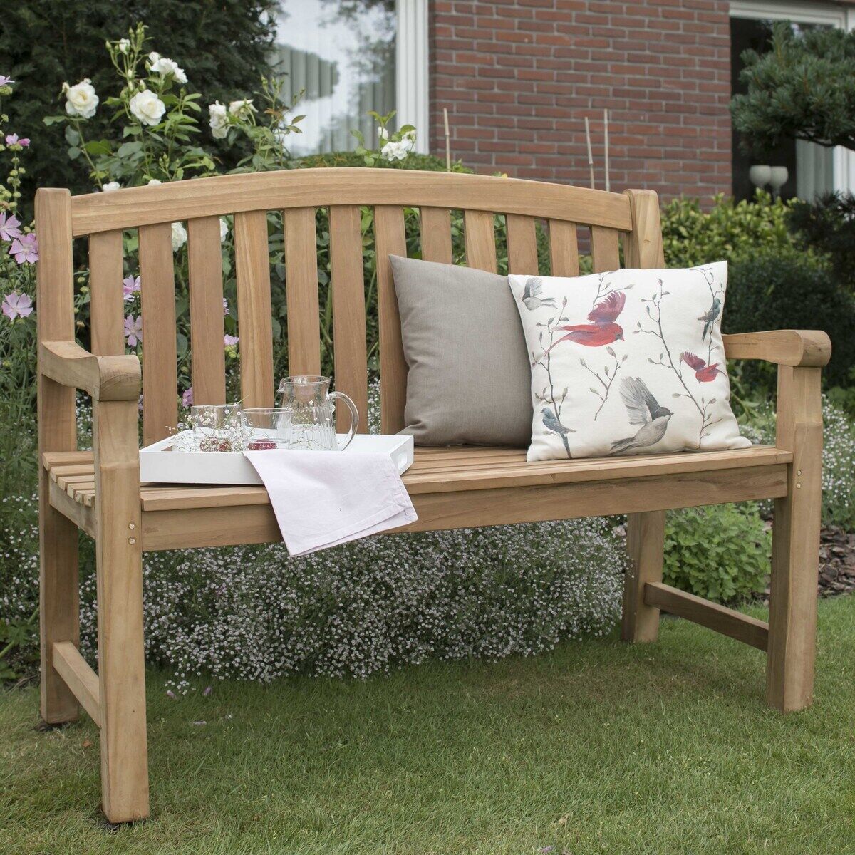 Auf einer Wiese im Garten steht eine helle Teakholz-Gartenbank »Swindon« von bene living mit zwei gemusterten Kissen, Tablett mit Glaskaraffe, Trinkglas und Serviette. Im Hintergrund blühende Blumen und ein Backsteinhaus mit Fenster.