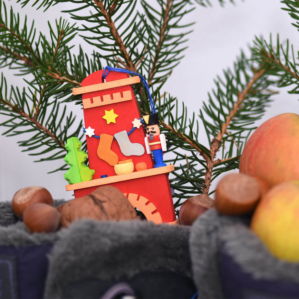 Handgefertigter weihnachtlicher Baumschmuck in Form eines Kamins mit Weihnachtsdekoration aus Holz, hängend an einem Tannenbaumzweig, umgeben von Nüssen und Äpfeln.
