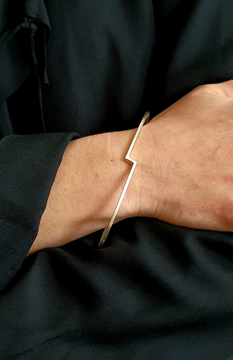 Schlanker, goldener Armreif mit einem zickzackförmigen Design, getragen am Handgelenk einer Person mit schwarzer Kleidung.