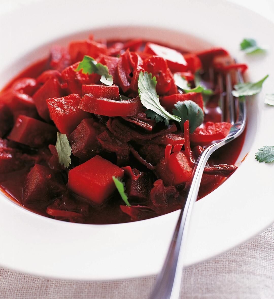 Nahaufnahme eines Tellers mit einem roten Eintopf oder Curry, garniert mit frischen grünen Kräutern und einer Gabel auf der rechten Seite. Der Eintopf enthält verschiedene Gemüsestücke und sieht köstlich und einladend aus.